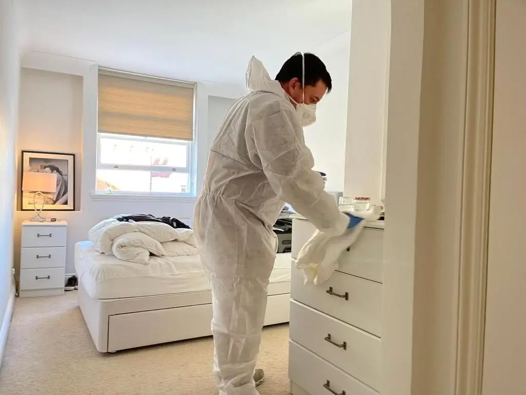 A cleaning technician in coveralls cleaning soot and ash from bedroom furniture.